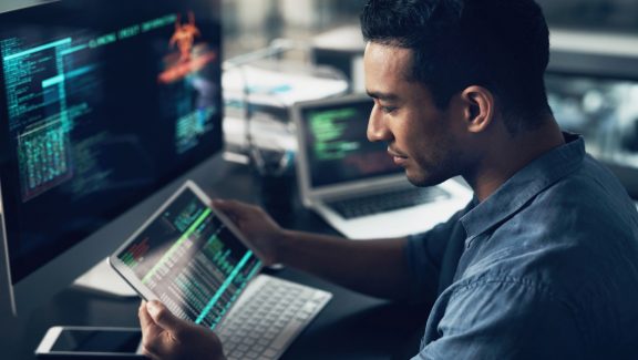 Man surrounded by computer screens looking at tablet